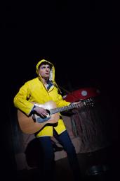 Orpheus plays a guitar, wearing a bright yellow raincoat